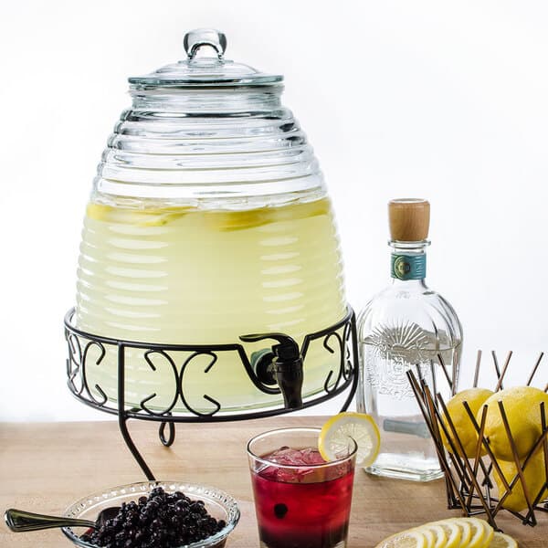 A glass jar with a drink and a black spigot filled with lemonade and berries with lemons on the table.