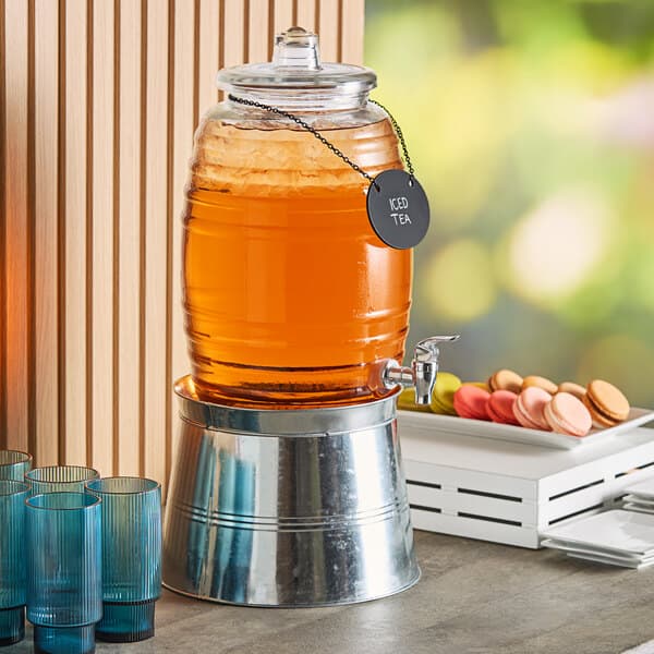 An Acopa glass beverage dispenser on a metal stand with a chalkboard sign filled with orange juice on a table.