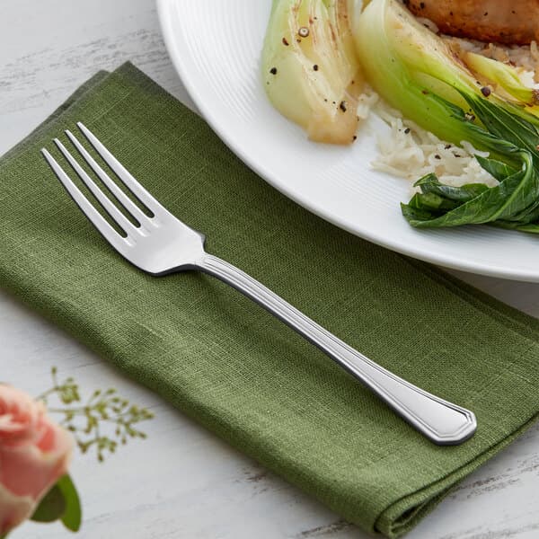 An Acopa stainless steel dinner fork on a napkin next to a plate of food.