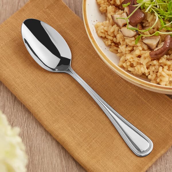An Acopa Edgeworth stainless steel serving spoon next to a bowl of rice with mushrooms and green sprouts on a table.