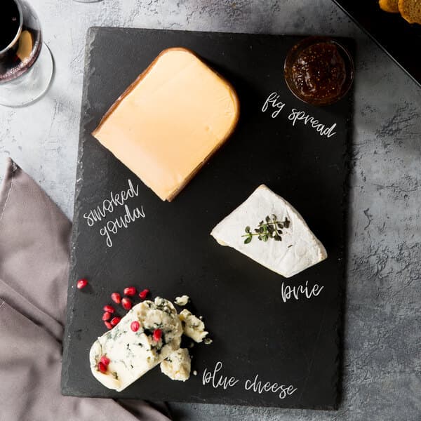 A black slate tray with pieces of cheese, wine, and crackers with a piece of blue cheese with pomegranate seeds on it.