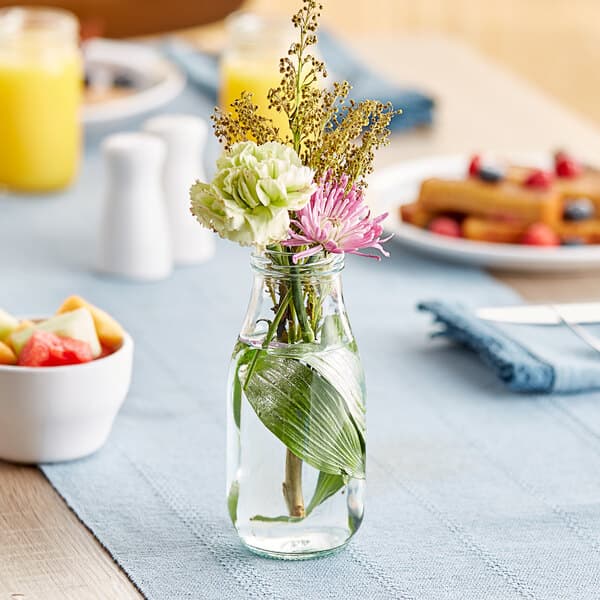 An Acopa glass milk bottle filled with flowers on a table with a bowl of fruit.