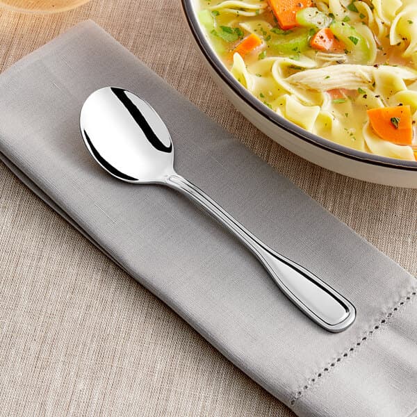 An Acopa Scottdale stainless steel bouillon spoon on a napkin next to a bowl of soup.