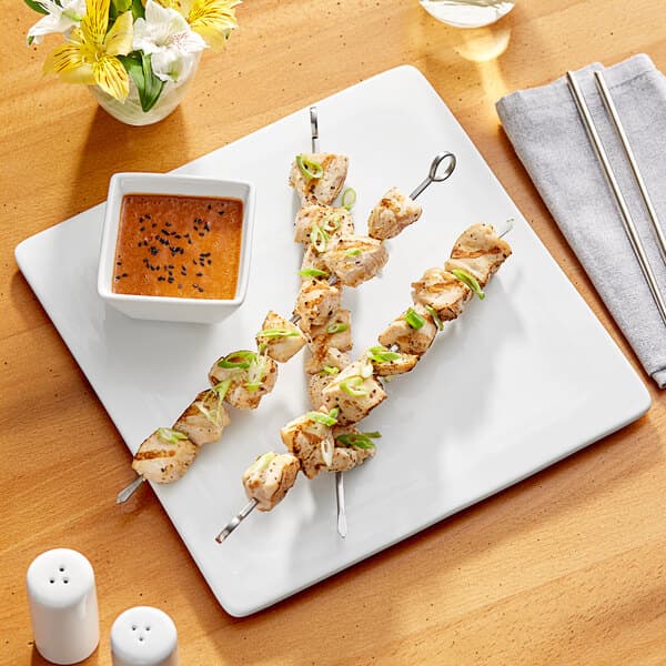 A plate of food with chicken skewers and dipping sauce on a white Acopa square plate.