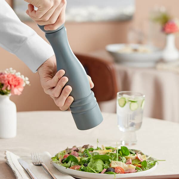 A hand using an Acopa steel blue wooden pepper mill to season a salad.