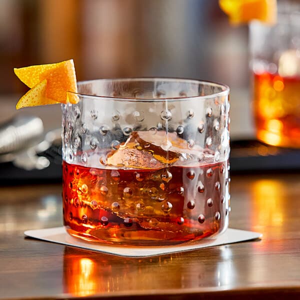 An Acopa hobnail style rocks glass with a drink, ice, and orange peel on a table.