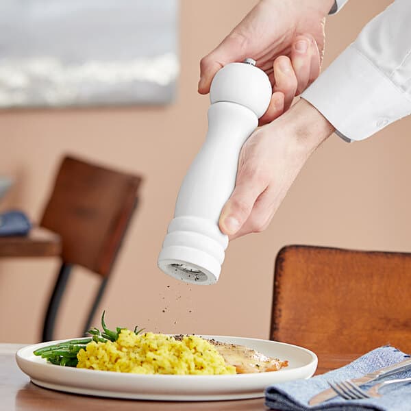 A person using a white wooden pepper mill to salt a plate of food.