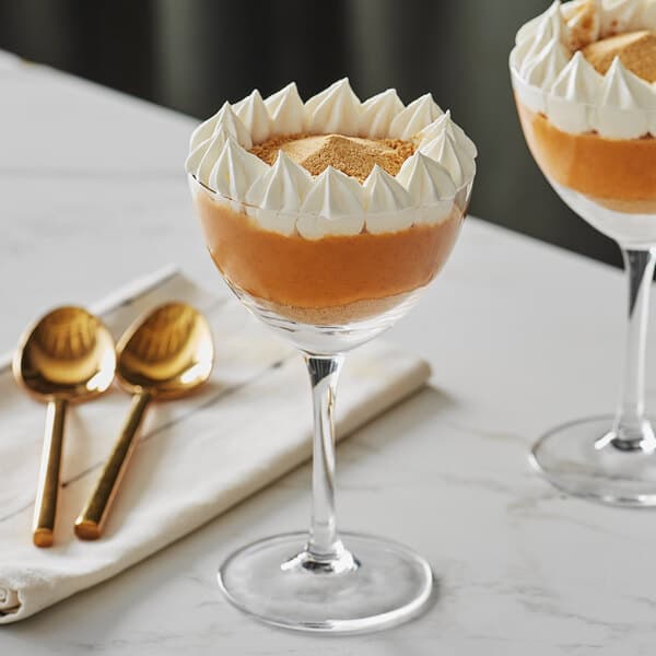Two Acopa Nick and Nora glasses filled with dessert and topped with whipped cream on a table with golden spoons.