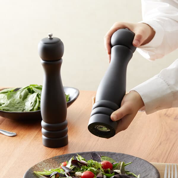 A person using a black Acopa wooden pepper mill to grind pepper over a salad.