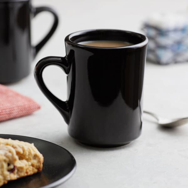 A black mug with a handle on a white surface with a plate of food and a black mug with a handle and a spoon.