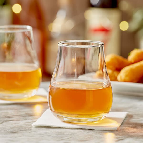 Two Acopa Select whiskey glasses filled with brown liquid on a table with food.