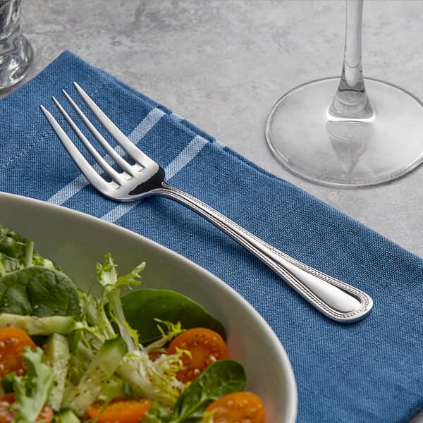 An Acopa Lydia stainless steel salad fork on a napkin next to a bowl of salad.