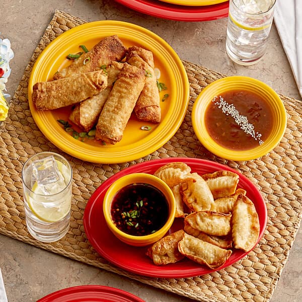 A table with Acopa Capri mango orange stoneware plates of fried spring rolls, dumplings, and food with a glass of water.
