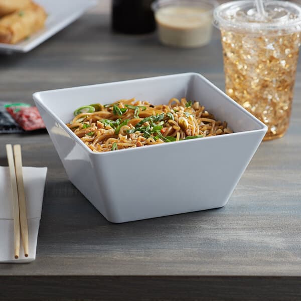 A bowl of noodles with green onions and nuts in an Acopa white melamine bowl on a table with a drink.