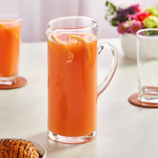 An Acopa slim glass pitcher of orange juice with lemon slices on a table.