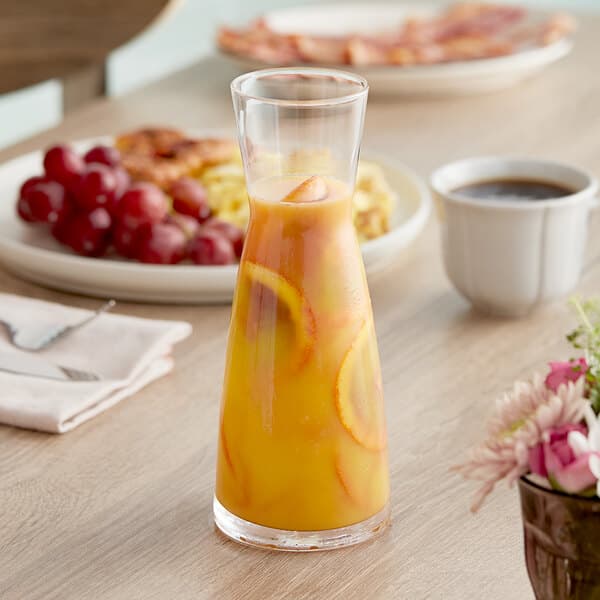 An Acopa glass carafe filled with orange juice on a table with fruit.