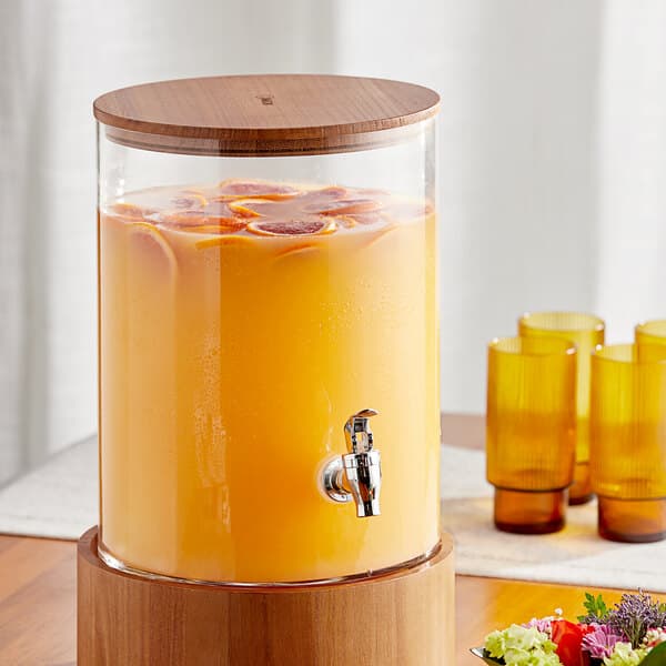 An Acopa glass beverage dispenser with orange liquid and a wooden lid.