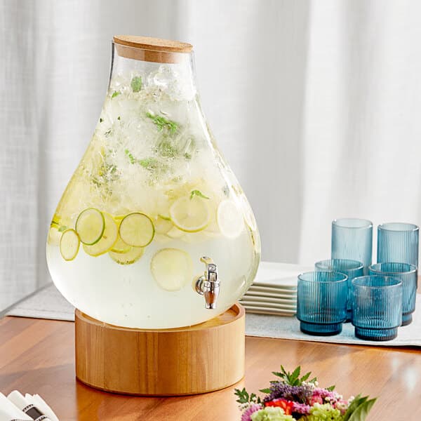 An Acopa glass beverage dispenser with liquid and lemon slices on a wood stand.