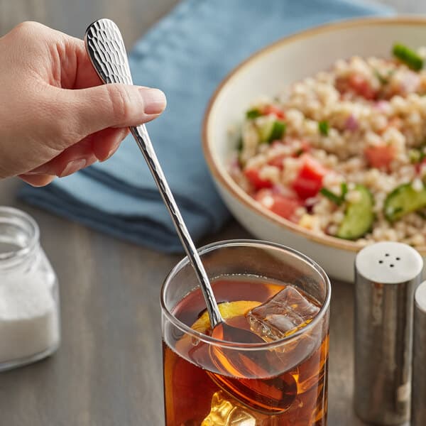 A hand holding an Acopa stainless steel iced tea spoon in a glass of liquid with ice and a bowl of rice.