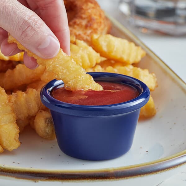 A person dipping a french fries into a blue Acopa melamine ramekin with ketchup.