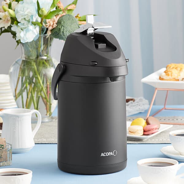 A black Acopa stainless steel coffee airpot on a table with a white cup of coffee.