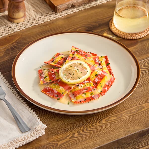 A round stoneware coupe plate with a vanilla bean color and a brown rim, holding striped ravioli garnished with a lemon slice and herbs.