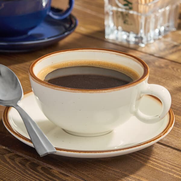 A cream-colored stoneware cup filled with black coffee, resting on a matching saucer with a spoon beside it.