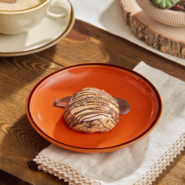A small orange stoneware coupe plate with a pastry drizzled in chocolate, placed on a wooden table.
