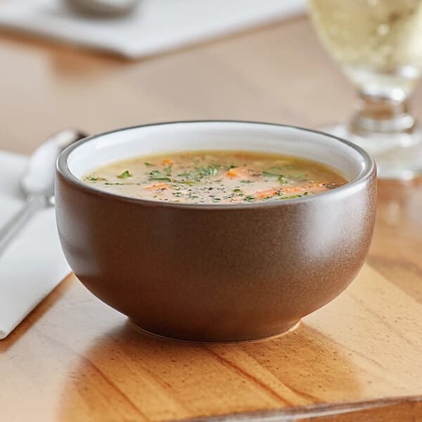 A brown matte stoneware bowl filled with soup, placed on a wooden table.