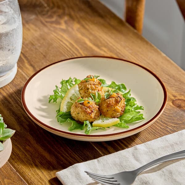 A round stoneware plate with a vanilla bean color and brown rim, holding three crab cakes on a bed of greens with lemon wedges.