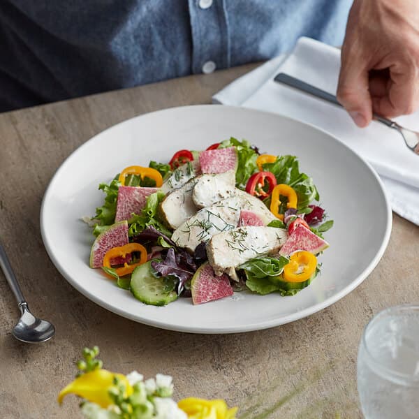 A hand holding a plate with a salad with chicken and vegetables on a white coupe plate.