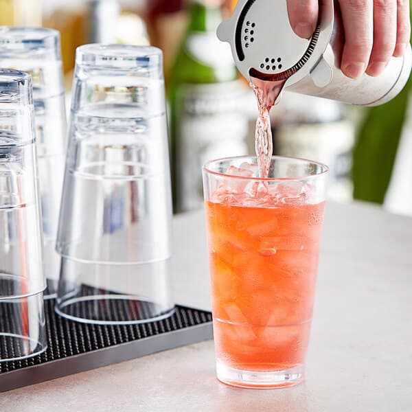 A hand pouring orange liquid from a green bottle into a Acopa Tritan plastic glass filled with ice.