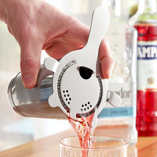 A hand using an Acopa silver Hawthorne strainer to pour a drink from a shaker into a glass.