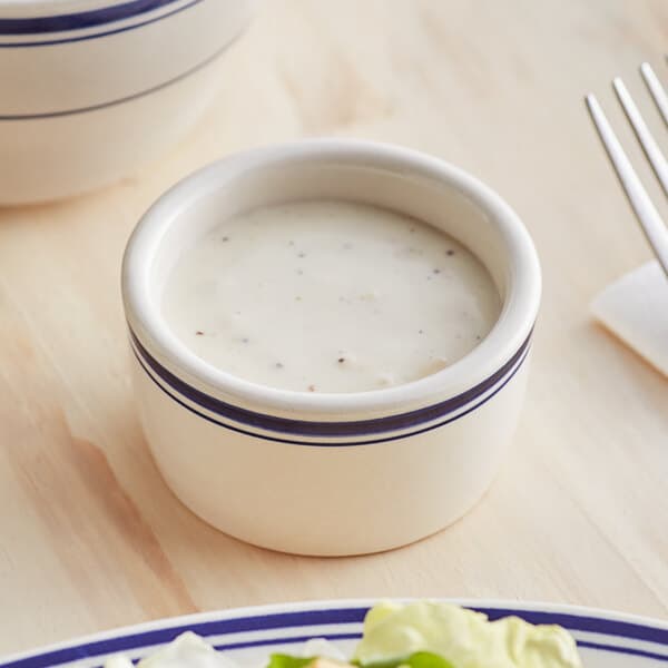 An Acopa ivory stoneware ramekin with a blue band filled with white sauce on a table.