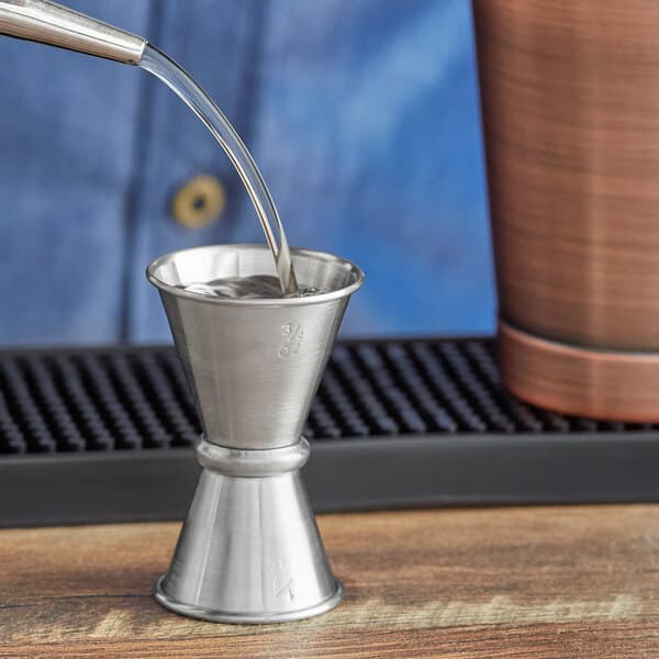 A person using an Acopa stainless steel Japanese jigger to pour liquid into a metal cup.