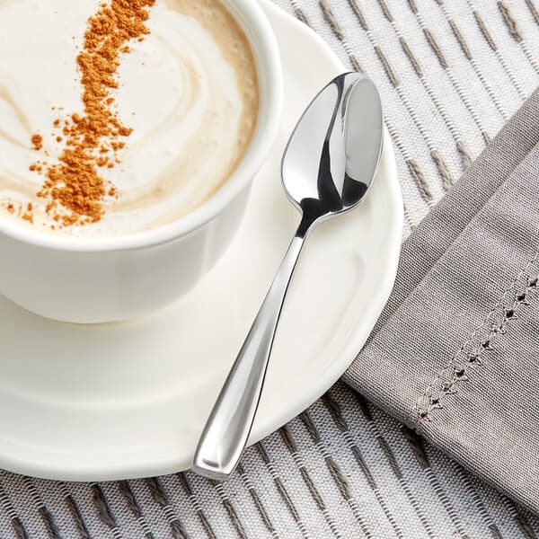 An Acopa stainless steel demitasse spoon in a cup of coffee with a swirl of brown powder on a saucer.