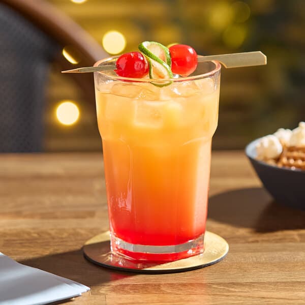 An Acopa Memphis beverage glass filled with ice, orange and red liquid, and a cherry on top on a coaster.