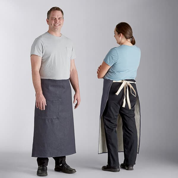 A man and woman standing next to each other wearing Acopa Kennett blue denim bistro aprons with natural webbing.