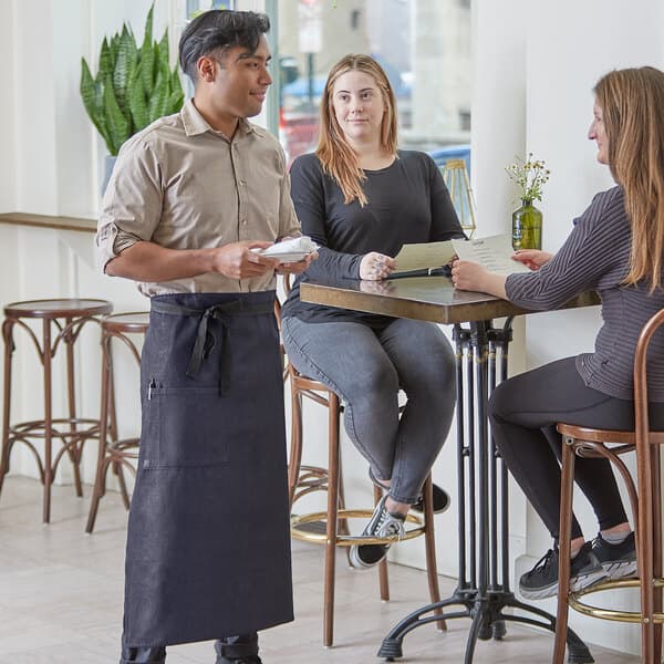 A man and woman sitting at a table in a farm-to-table restaurant with a waiter wearing a blue denim Acopa Kennett Bistro apron.