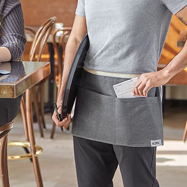 A man wearing a gray Acopa Kennett denim waist apron with natural webbing holding a black folder and a pen at a table.