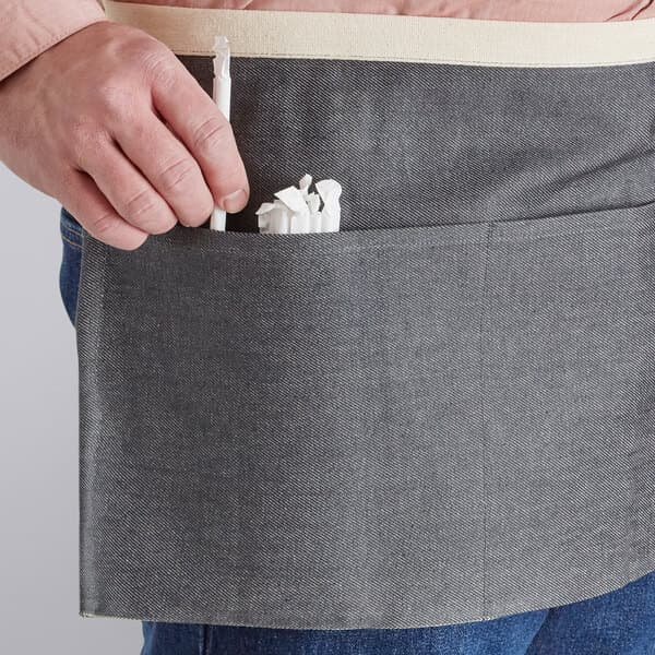 A person's hand holding a white object in a gray Acopa Kennett waist apron with natural webbing and three pockets.
