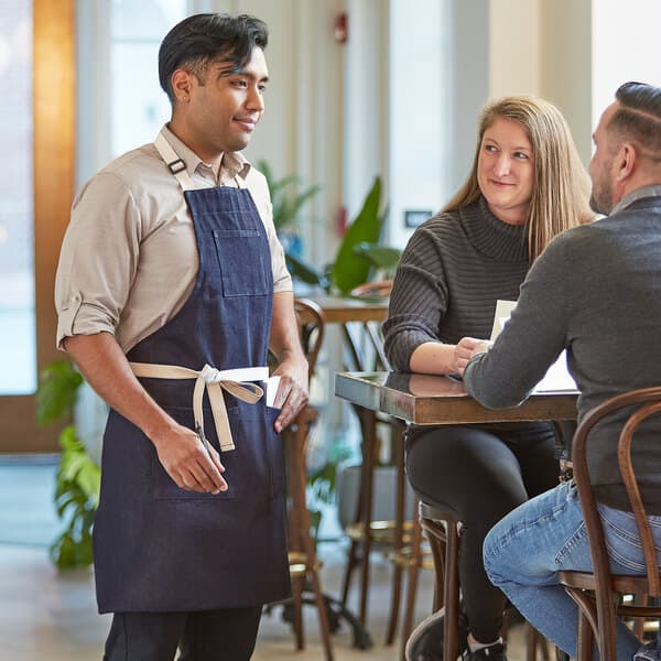 A man in a blue Acopa Kennett apron talking to a couple at a farm-to-table restaurant.