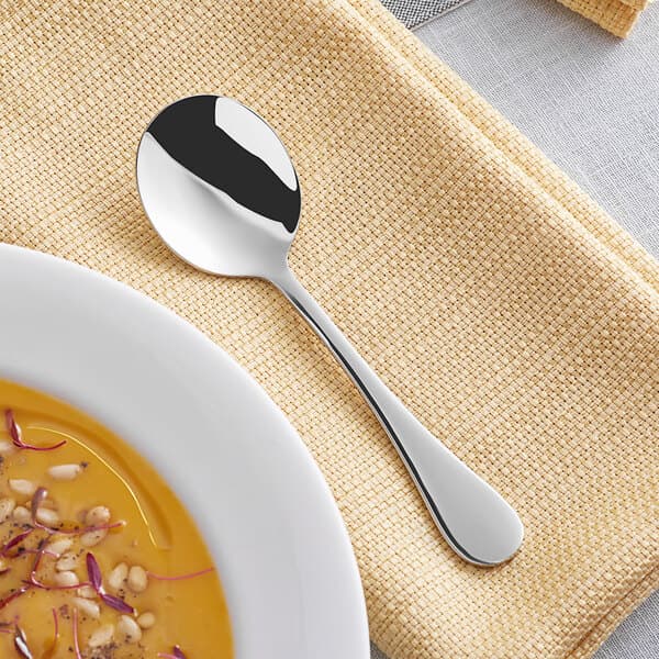 An Acopa Vittoria stainless steel bouillon spoon on a plate with a bowl of soup.