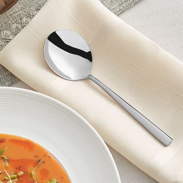 An Acopa stainless steel bouillon spoon on a napkin next to a bowl of soup.