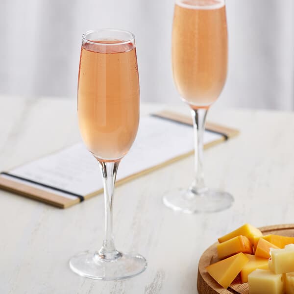 Two Acopa flute glasses of pink liquid next to a plate of cheese on a wood surface.