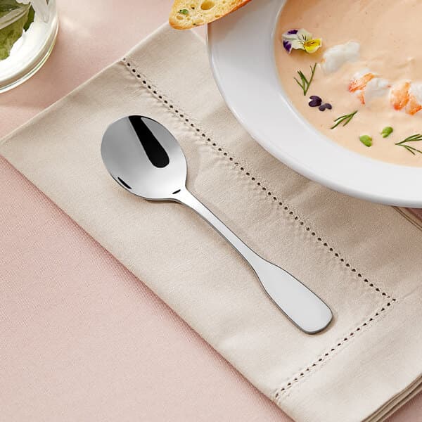 An Acopa stainless steel bouillon spoon on a napkin next to a bowl of soup.