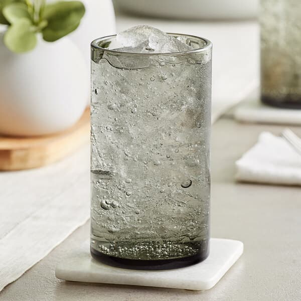 A close-up of a Acopa Pangea beverage glass with ice water on a coaster on a table with a plant.