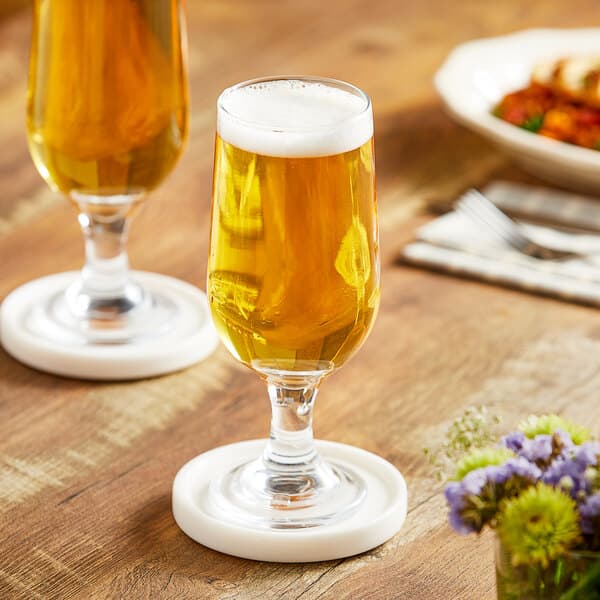 Two Acopa Select stemmed pilsner glasses of beer on coasters on a wooden table.