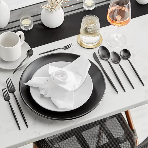 A table set with a white tablecloth, black plates, and Acopa Odin brushed stainless steel forks.