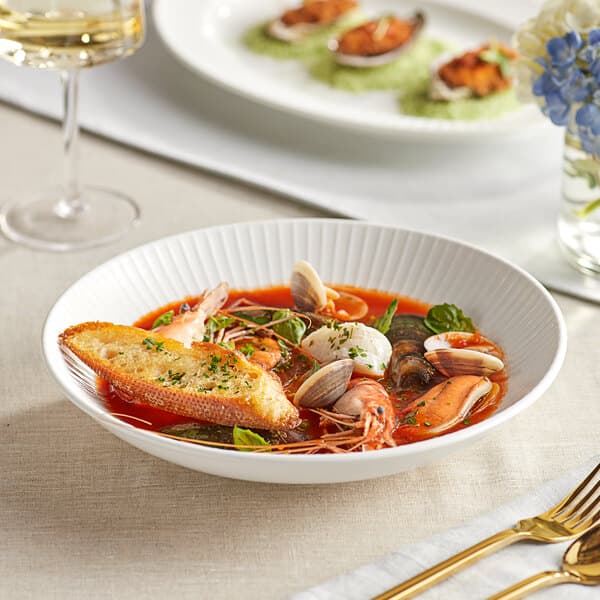 A bowl of Acopa Cordelia porcelain soup with seafood and bread on a white background.